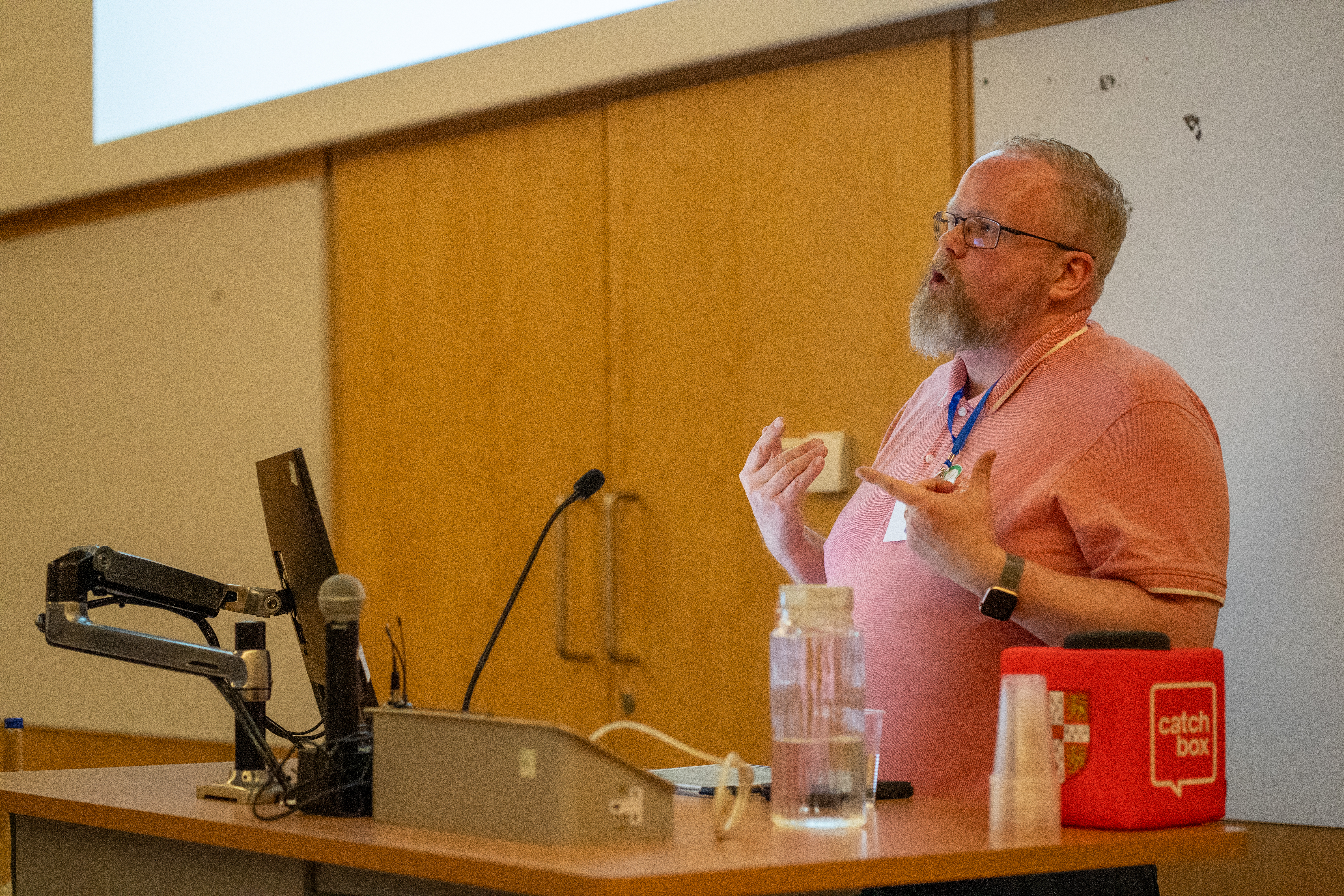 Man in pink shirt speaking at podium with microphone and laptop