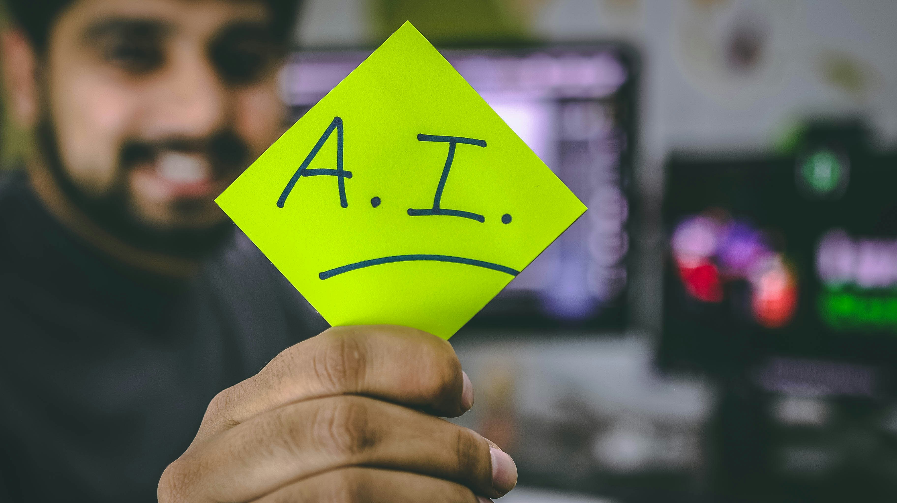 Person holding bright yellow sticky note with "A.I." written on it in front of computer screen